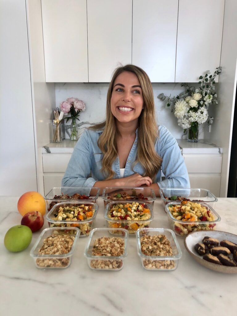 Lyndi Cohen in her kitchen with 9 full food containers, fresh fruit and a big smile.